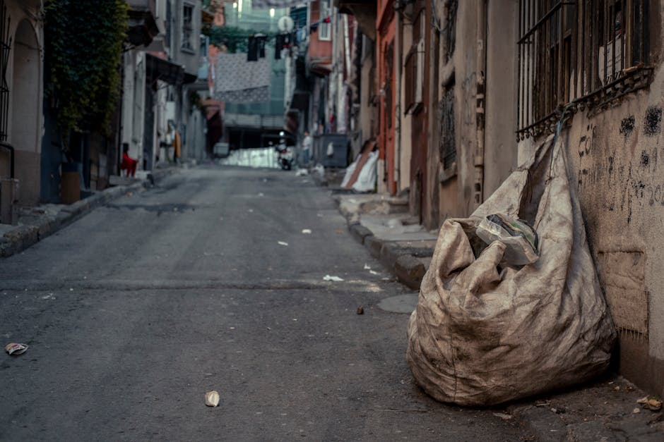 An outdoor scene on a paved urban street showing overflowing waste and rubbish bags piled around large communal rubbish skips. The waste includes a mix of cardboard boxes, plastic bags, and paper debr