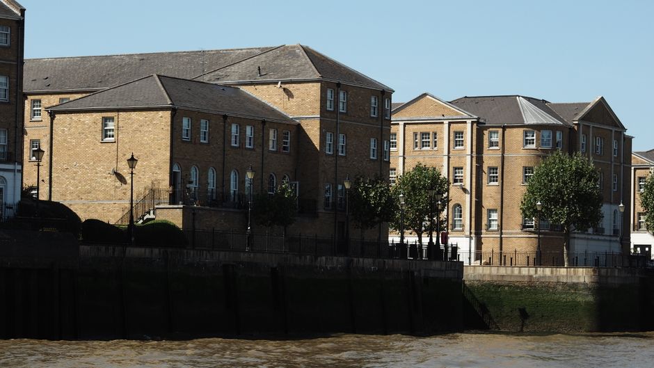 A row of multi-storey brick residential buildings situated along a riverside, with several small, neatly trimmed trees planted in front of them. The buildings feature uniform brownish-yellow brick fac